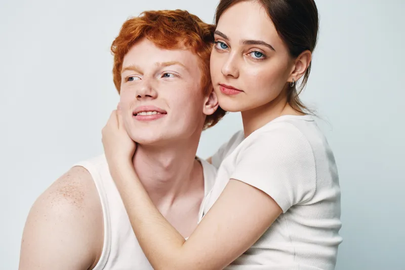 Young couple with red hair and blue eyes in casual clothing embracing and posing at studio with neutral background for love and relationship concept.