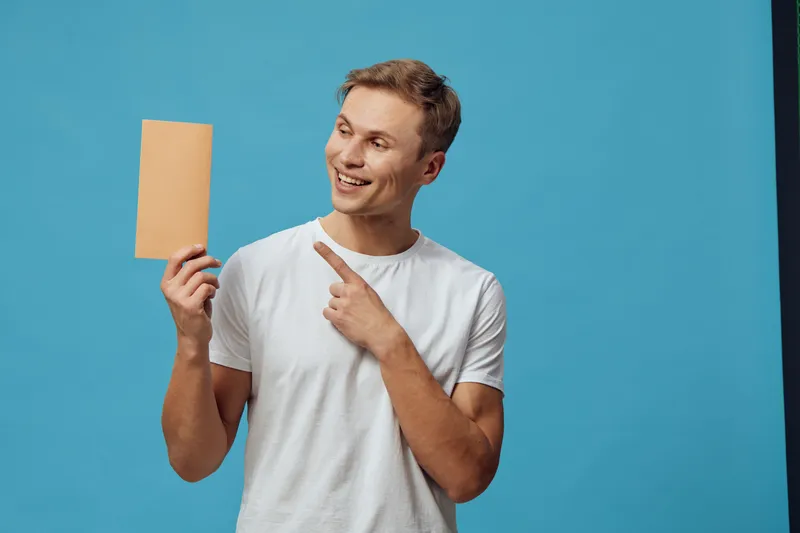 Smiling young man with short hair holding a blank cardboard sign in front of a blue background. Person wearing casual white t-shirt, expressing excitement and friendliness. Studio portrait, lifestyle 