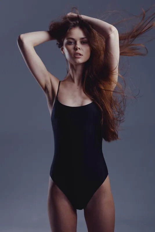 Young woman with flowing hair posing in black swimsuit with confident expression against dark background