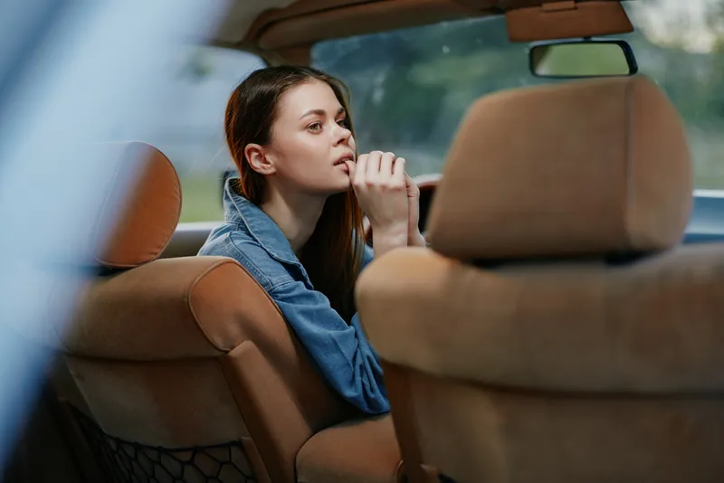 Young woman contemplating in the car with a pensive expression, wearing a denim jacket and set against a blurred nature background, evoking feelings of solitude and reflection.