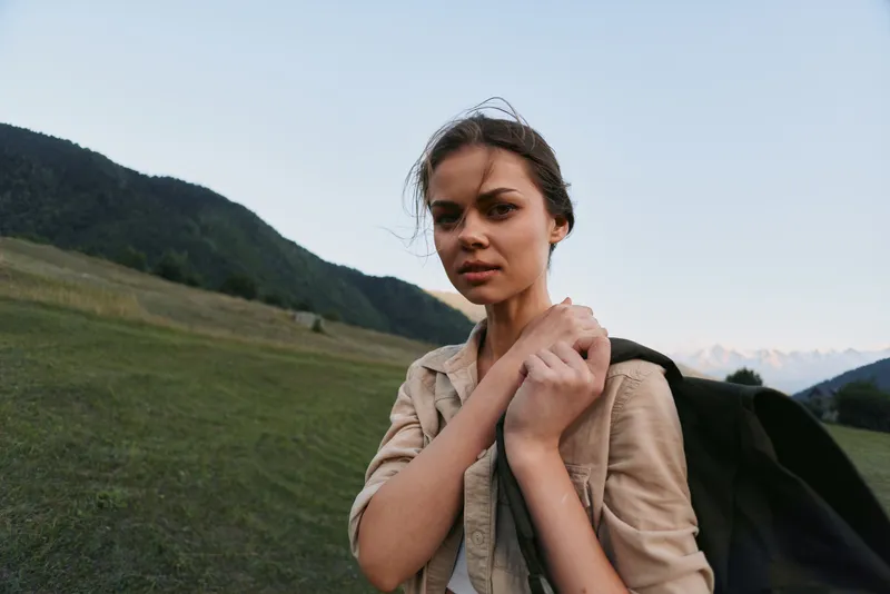 Young woman in casual clothes stands outdoors in a mountainous area with green hills behind her, looking confidently at the camera with natural light.