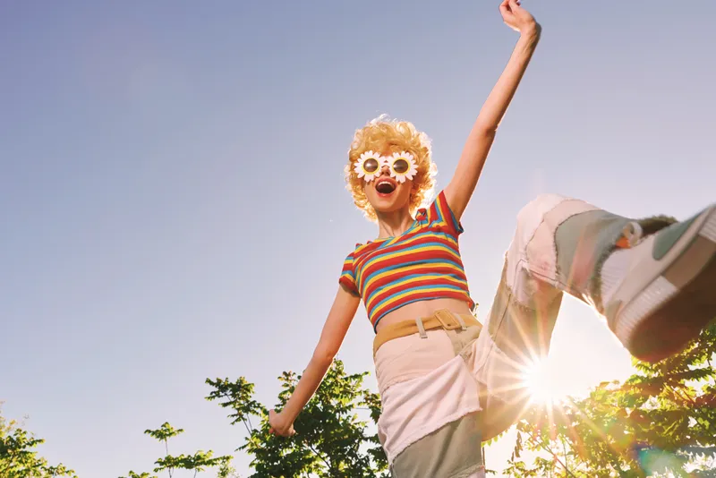 Joyful woman outdoors wearing a striped top and light shorts, floral sunglasses, arms raised, sun flare behind. Retro color film vibe, playful lifestyle moment, fashion.