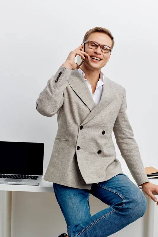smiling man talking smartphone wearing casual jacket glasses sitting white desk laptop digital nomad remote work concept