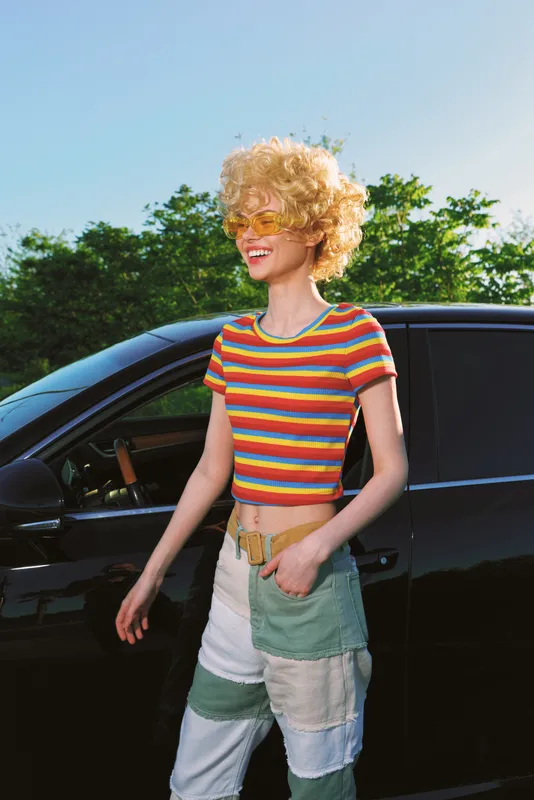 Smiling woman in a rainbow striped crop top stands beside a car outdoors, wearing patchwork denim. Retro film color and warm lighting convey lifestyle, fashion, and carefree summer mood.
