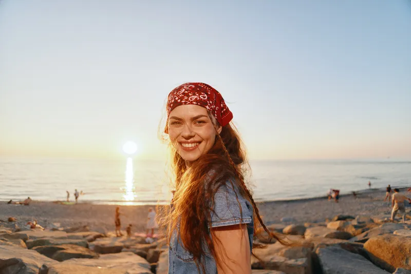 lifestyle woman in modern boho-western denim street style smiles warmly on rocky beach at sunset with film color tones creating relaxed vibe
