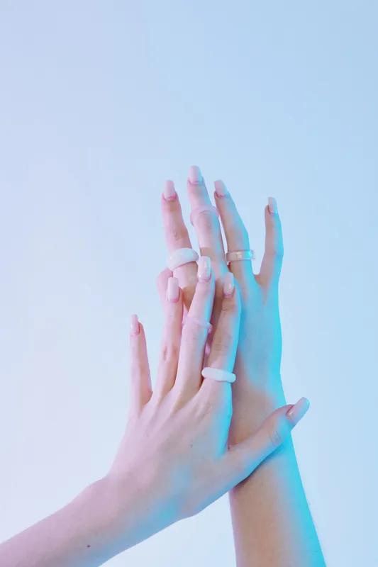 Close-up of two female hands with manicured nails and multiple rings. Soft blue and pink lighting highlights the delicate skin and beauty accessories. Concept of fashion and elegance.