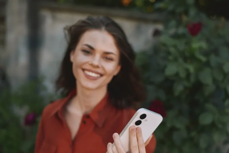 Vibrant woman outdoors, smiling into a smartphone selfie, casual outfit, sunlit garden setting, relaxed pose, natural colors, fresh breeze, candid lifestyle moment, luminous joy
