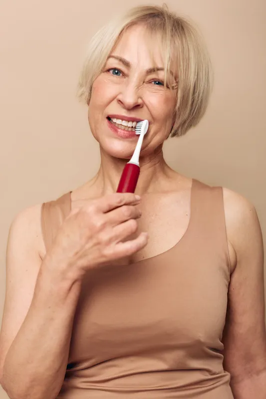 Mature woman with short blond hair brushing teeth with electric toothbrush. Close-up portrait on beige background. Concept of dental hygiene, oral care, health routine, and senior wellness.