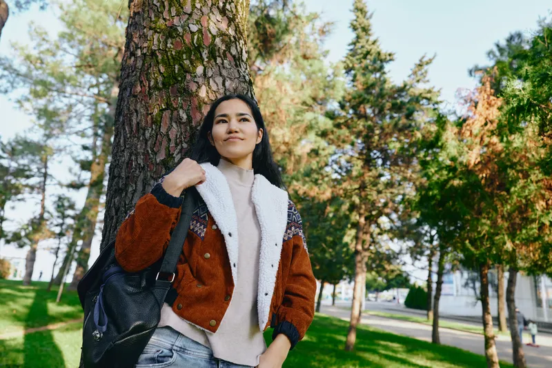 Woman with backpack leaning near a tall tree in a sunlit park, autumn colors, casual fashion, outdoor scene, relaxed mood, nature backdrop for lifestyle and fashion stock