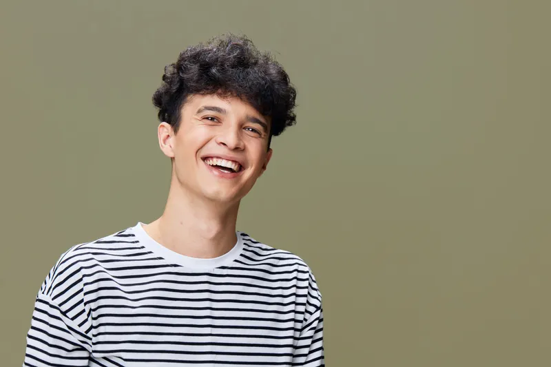 smiling man wearing striped shirt with curly hair, joyful expression on beige background, casual portrait with natural lighting and relaxed mood