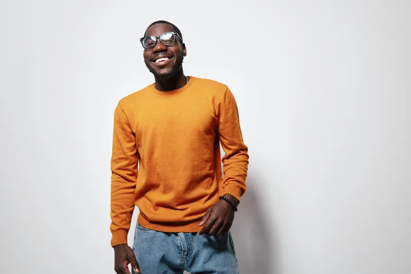 African American man with dark skin wearing glasses and orange sweater smiling confidently against white background. Casual style, happy expression, standing and posing for portrait.