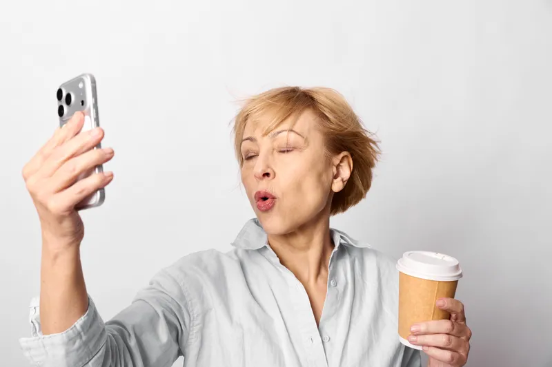 Surprised elderly woman holding smartphone and coffee cup indoors portrait on clean light background, using communication with digital device, casual lifestyle, soft studio lighting