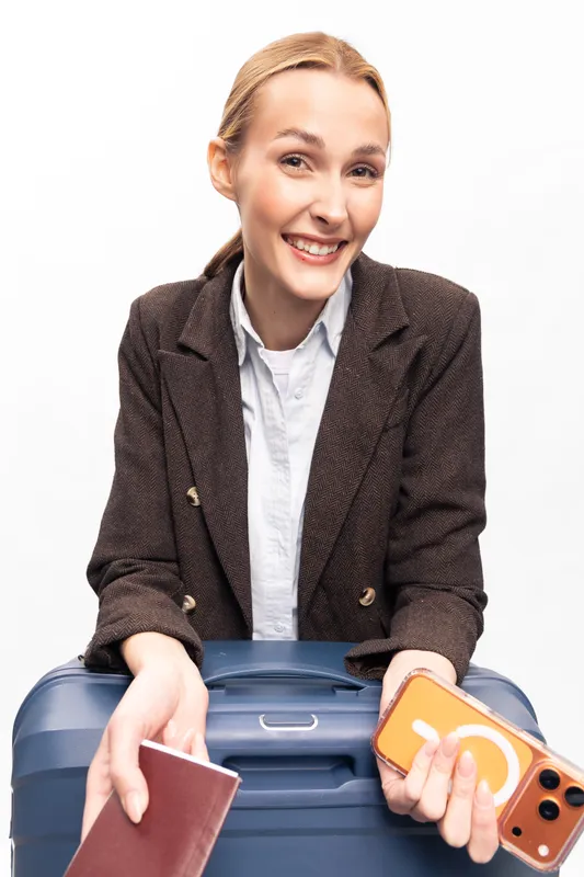 Smiling young woman in brown blazer holds a passport and smartphone while leaning on blue suitcase. Travel concept with happy female traveler preparing for a trip against white background.