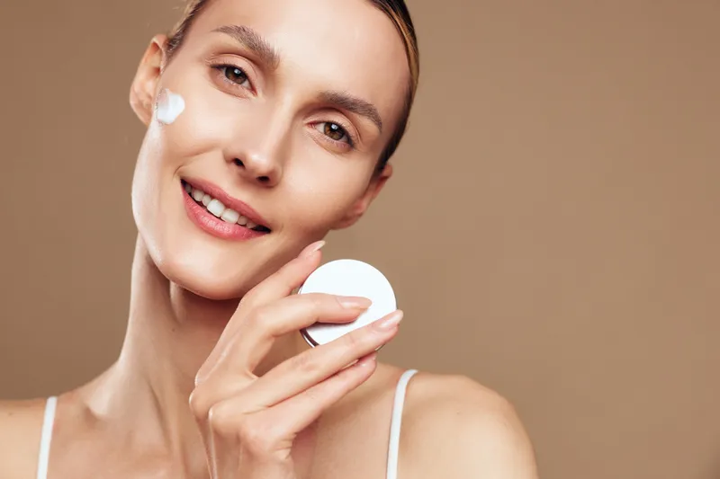 Smiling woman with clear skin applying white face cream on cheek using cotton pad. Close-up on beige background. Concept of skincare, beauty, self-care, and wellness with soft lighting.