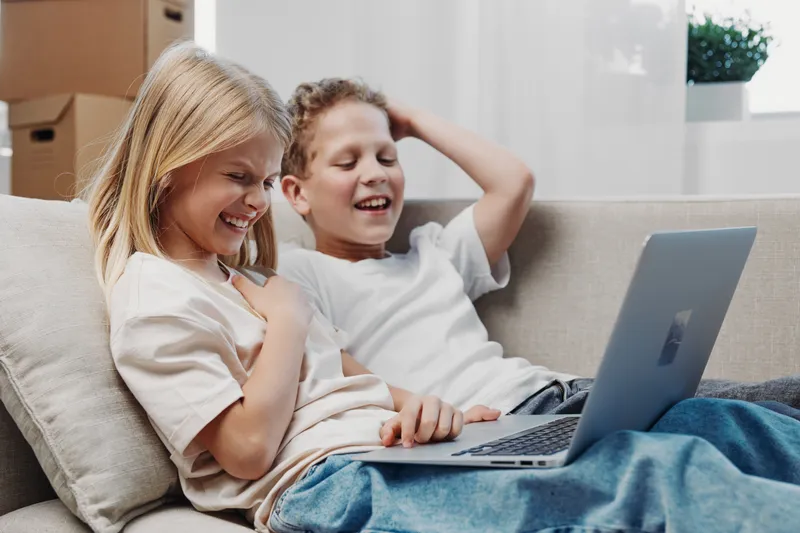 Two happy children sitting on sofa using laptop, laughing and enjoying digital entertainment at home in bright modern living room.
