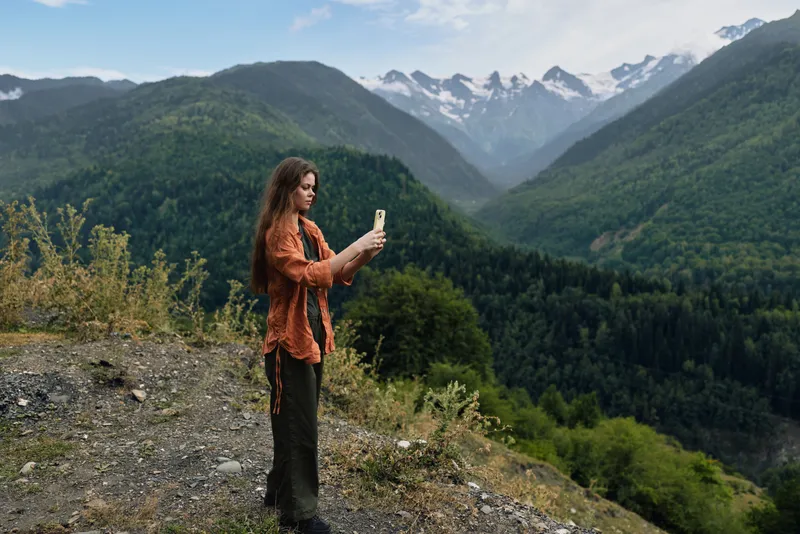 Woman standing on a hillside taking a selfie with a phone in a mountainous landscape, lush valley and distant snowy peaks, conveying a serene and adventurous outdoor moment.