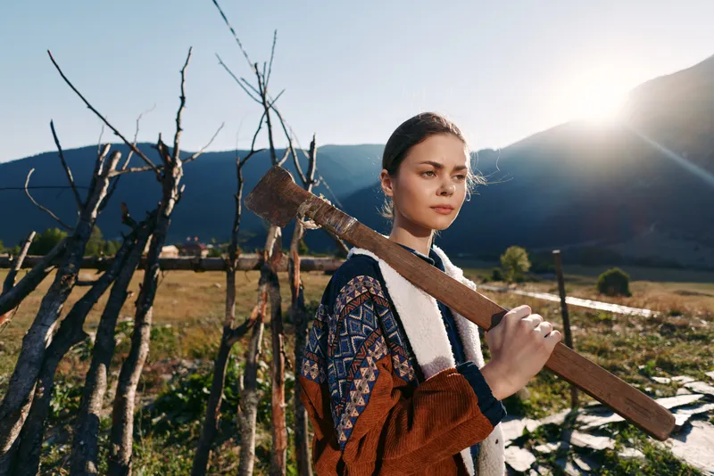 Woman with axe stands in rural field by wooden fence against mountains, portrait of determined farmworker in sunlight, traditional clothing and rustic landscape.