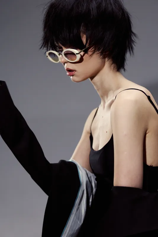 Stylish woman wearing sunglasses and black outfit with modern haircut in studio with neutral background.