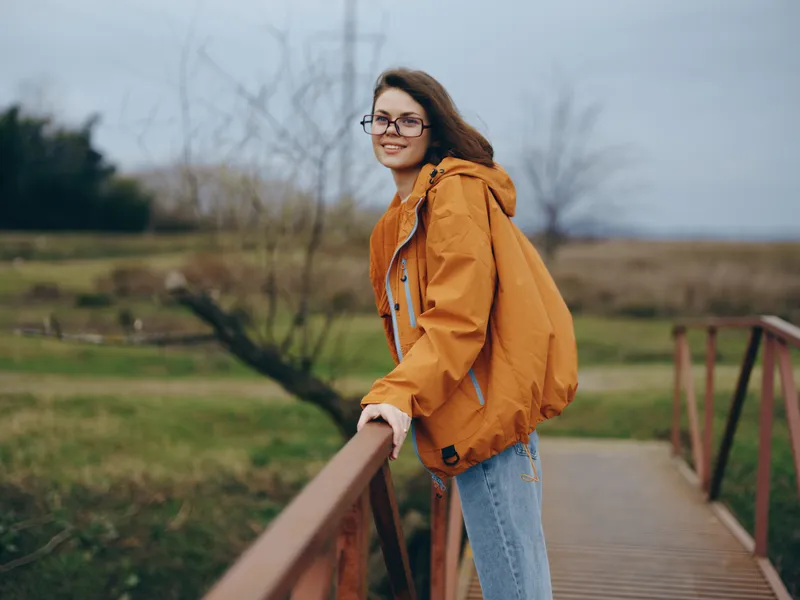 Woman outdoors lifestyle, orange jacket, denim jeans, glasses smile, standing on wooden bridge, rural park landscape under cloudy sky, casual travel mood, natural light