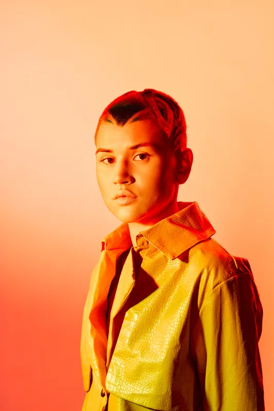 young woman with braided hair wearing a textured yellow jacket poses confidently against warm orange background, showcasing bold fashion and modern style.