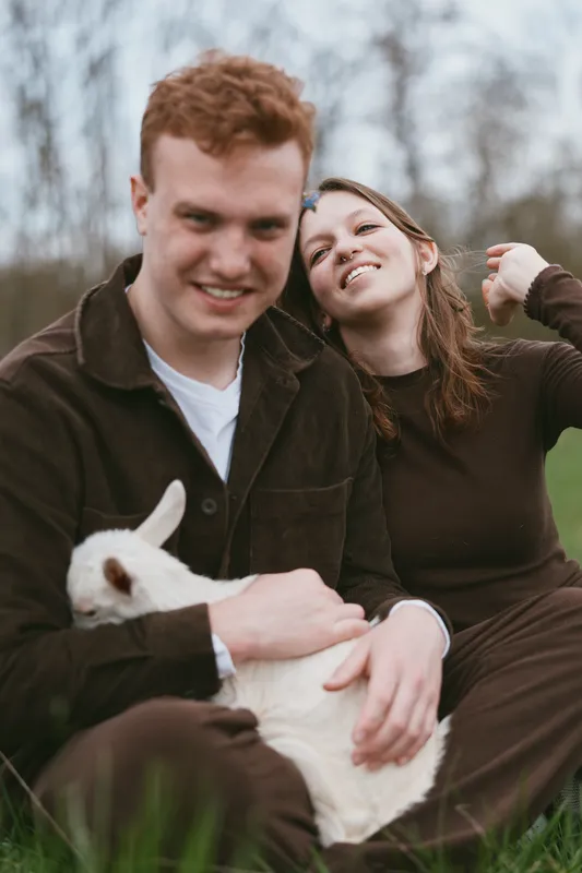 Couple outdoors with a playful goat kid, in a sunlit meadow, sharing laughter and warmth, a relaxed lifestyle moment, natural colors, candid connection, rustic countryside backdrop