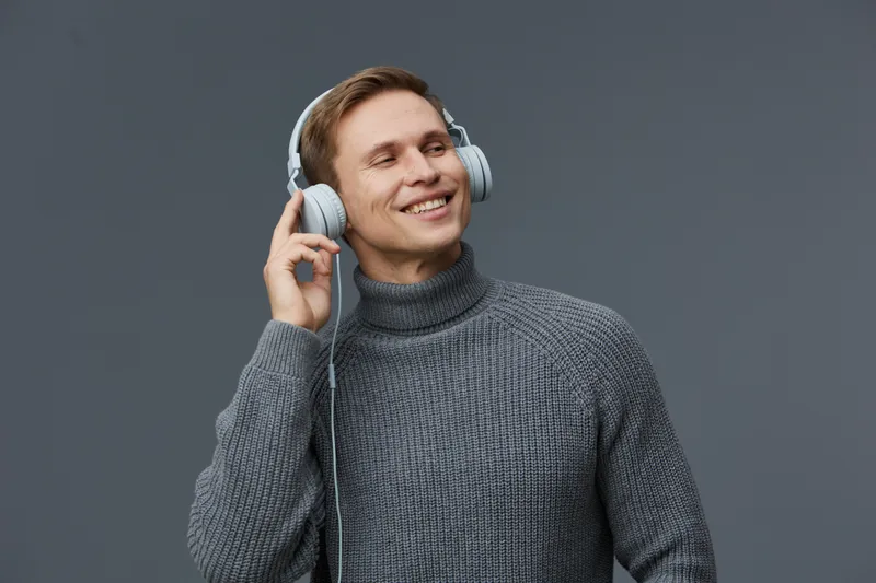 Smiling man listening to music with headphones on gray background. Portrait of a happy young adult male enjoying music, casual style with gray sweater. Concept: leisure, relaxation, entertainment.