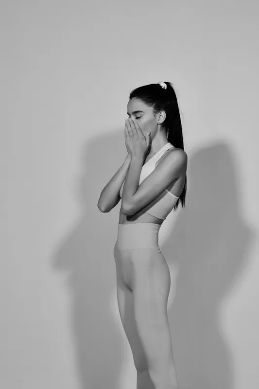 Black and white portrait of a young woman sport fitness athlete covering her face with hands, showing emotion and strength in minimal workout clothing, studio background.