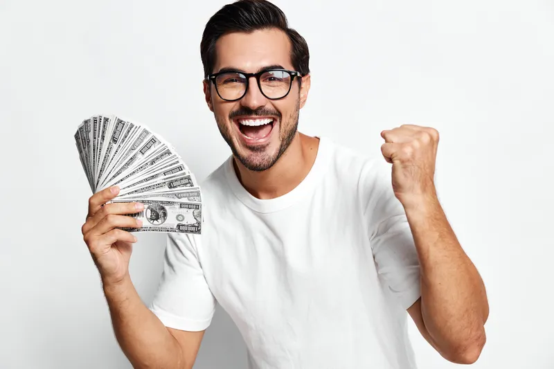 Happy smiling man holding a fan of cash with excitement, celebrating a financial win, wearing casual white t-shirt and stylish glasses, isolated on plain background.