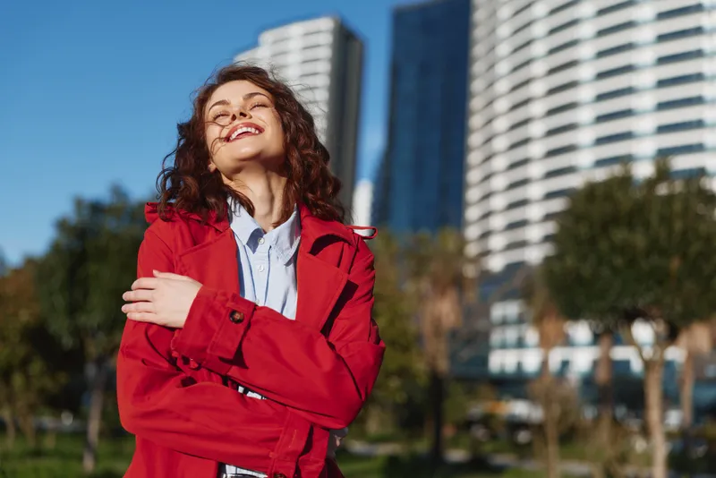 Young woman with curly hair wearing a red coat smiles joyfully with her eyes closed while standing outdoors in front of modern buildings on a sunny day.