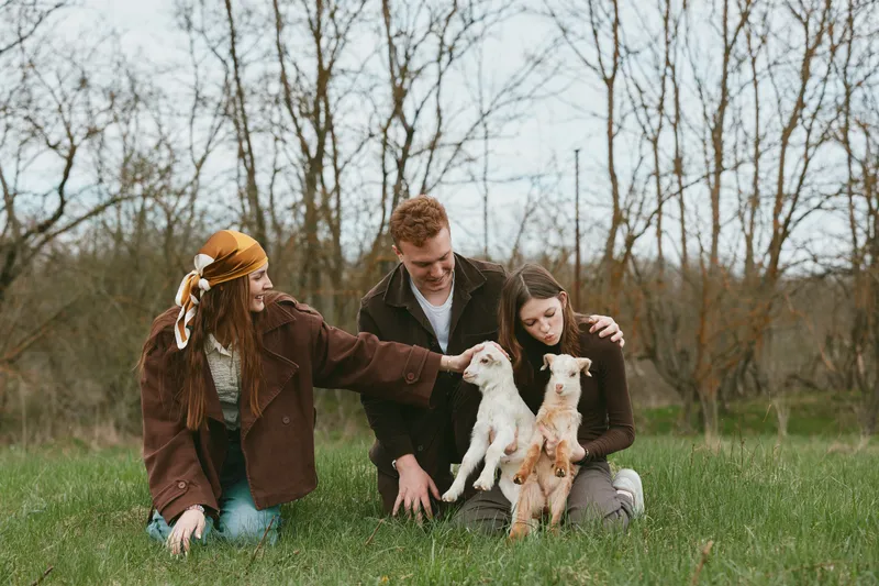 Group of friends in a park gently interact with goat kids, a warm lifestyle moment outdoors, celebrating nature, companionship, and playful animal encounters under soft daylight.