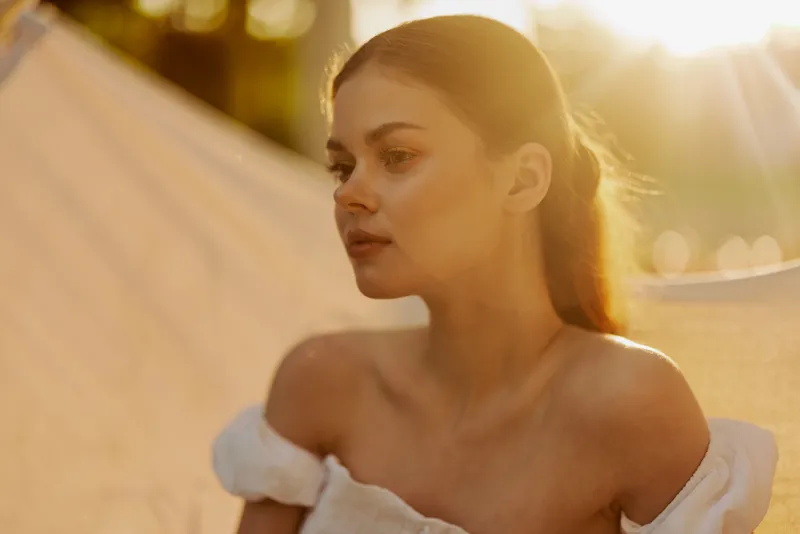 A serene young woman with long brown hair, wearing a white off shoulder dress, gazing thoughtfully during golden hour, capturing tranquility and beauty