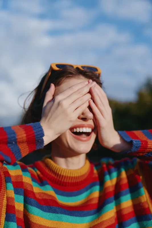 woman smiling striped sweater sunglasses covering eyes outdoor young adult joyful: Young adult woman smiling in a colorful striped sweater with sunglasses on head, covering eyes with hands while