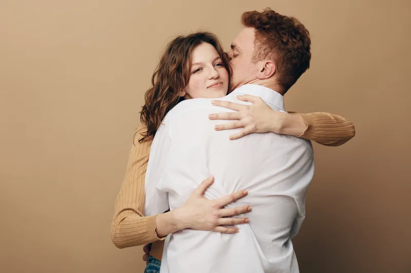 Young couple embraces warmly against beige background. Woman smiles softly while man kisses her cheek. Concept of love, affection, intimacy, and romantic relationship between partners.