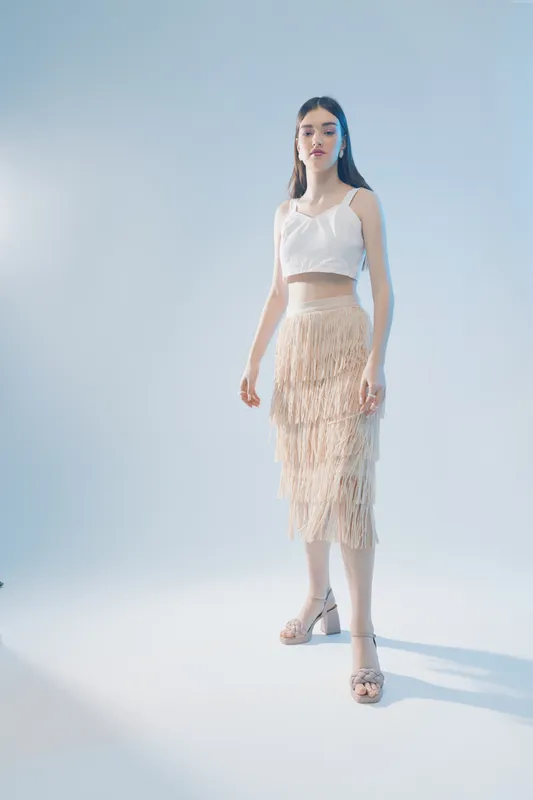 Young woman in white top and beige fringed skirt poses confidently in a studio with soft lighting. Concept of fashion, beauty, elegance, and modern style for lifestyle and clothing.