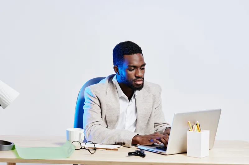 Professional young African American man in casual attire working on laptop at desk with office supplies and coffee mug in bright modern workspace, focused on task, isolated on plain white background. 