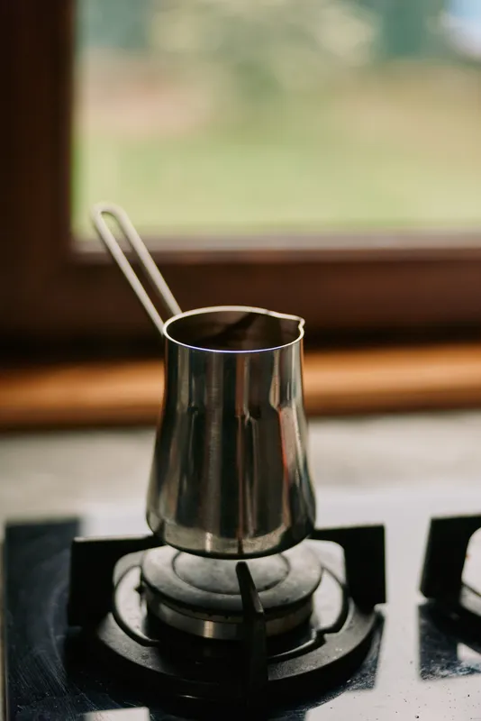 A small metal coffee pot sits on a gas stove burner in a cozy kitchen, steam rising as brewing begins, highlighting a warm homely cooking moment