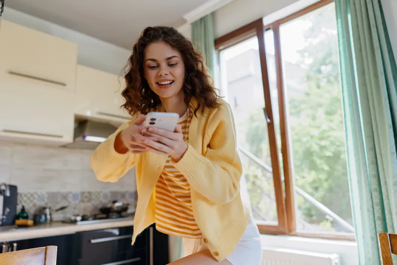 Smiling woman with curly hair wearing a yellow cardigan checks her smartphone in a bright home kitchen, natural light from a large window. Casual everyday lifestyle, cozy mood, warm colors.