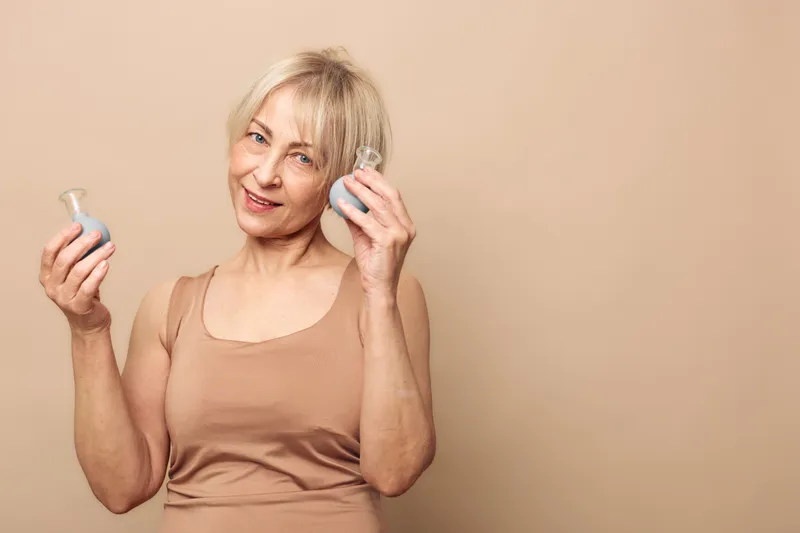 Smiling mature woman with short blonde hair holds two glass vases, wearing beige tank top. Neutral background highlights natural beauty and calm confident mood.
