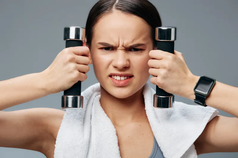 Studio portrait of a woman fitness, sport athlete lifting chrome dumbbells with towel on shoulders, gritty focused expression, visible sweat, athleticwear and strength training concept.