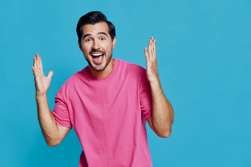 Smiling man in bright pink shirt joyfully raising hands, expressing excitement and happiness, posing against a solid blue background in a cheerful studio portrait.