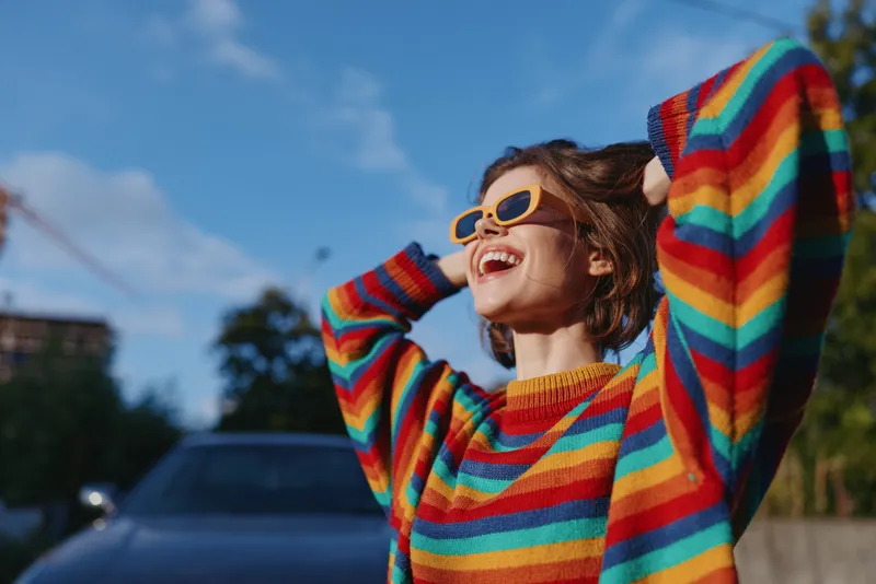 Woman smiling in rainbow sweater and sunglasses, young adult laughing with hands in hair outdoors by parked car in sunny urban setting, joyful lifestyle fashion travel vibe