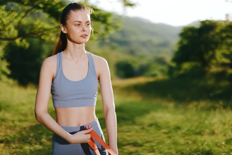 Athletic young woman in activewear holding resistance bands outdoors, showcasing strength and determination in a lush green environment