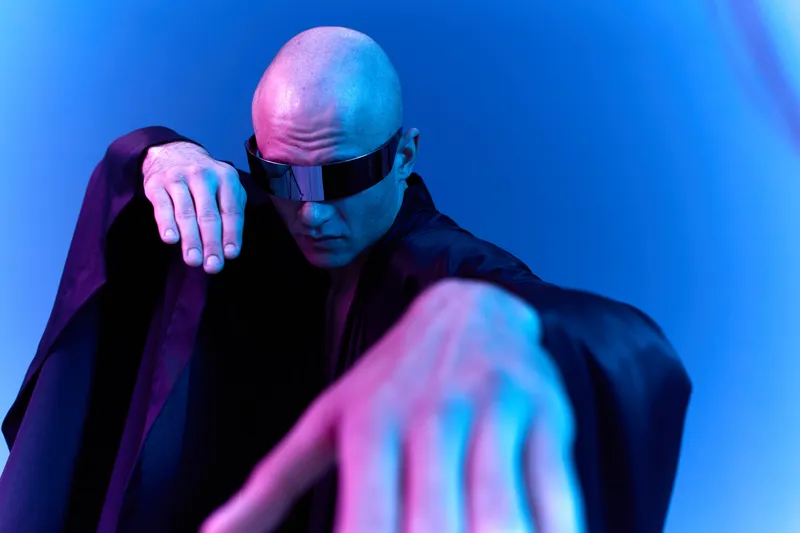 A striking bald man with sunglasses poses dramatically against a vibrant blue gradient background, showcasing futuristic fashion and a bold demeanor.