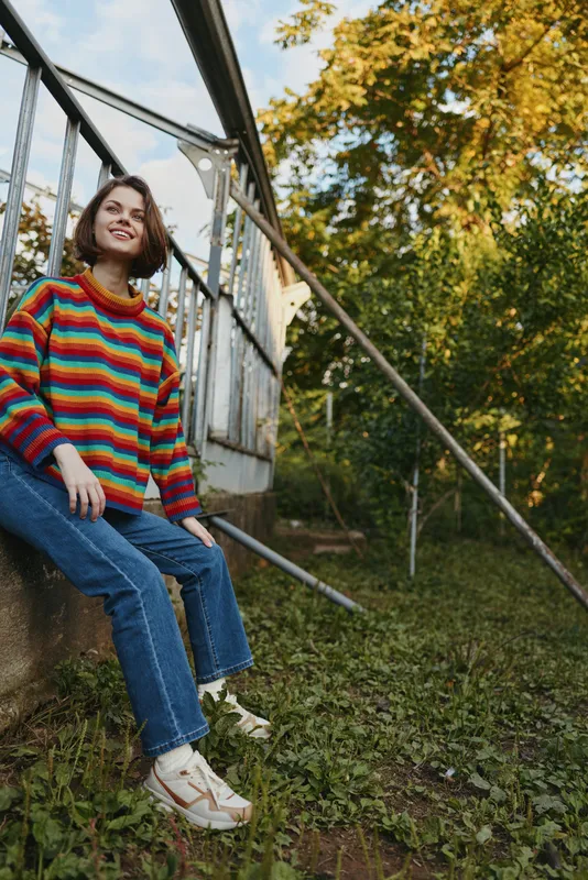 young woman in her mid 20s wearing a colorful striped sweater and jeans, sitting on backyard steps smiling and looking up, casual outfit, relaxed autumn lifestyle scene