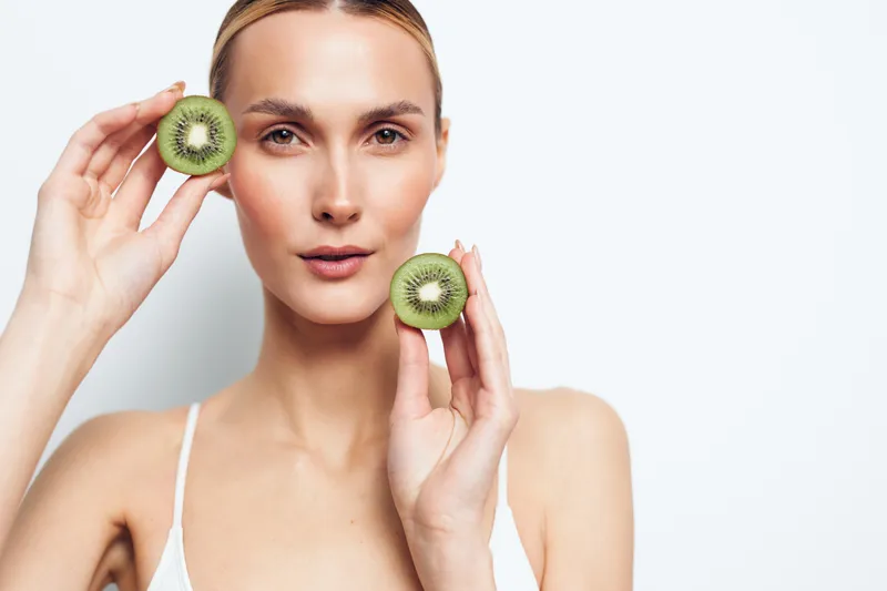 Young woman holds two halves of fresh kiwi fruit near face with calm expression isolated on white background closeup portrait of healthy lifestyle and natural beauty concept.