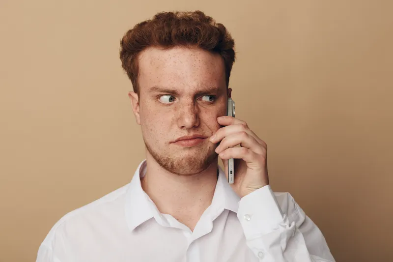 Young man with red hair and freckles wearing white shirt looks suspicious while holding smartphone to ear. Neutral beige background. Concept of doubt, communication, and uncertainty.