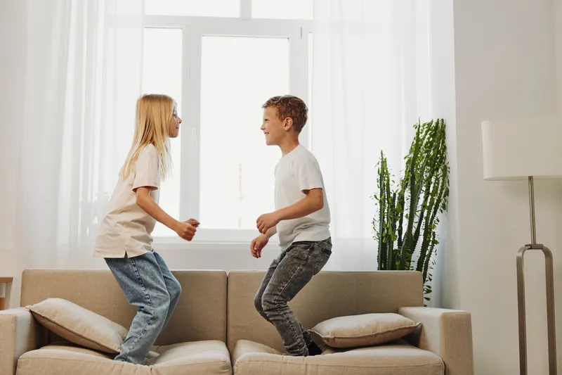 Two happy children jump on a beige couch in a bright living room with natural light. The boy and girl enjoy playful moments together, showing joy and carefree energy in a home environment.