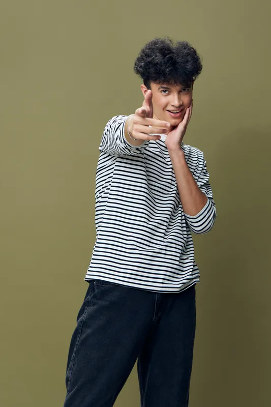 Teen boy wearing striped shirt pointing fingers with playful smile against olive background, curly hair, casual style, youthful energy and confident expression.
