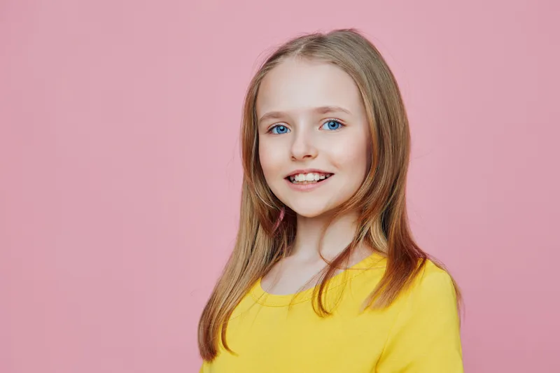 Smiling young girl with long blonde hair in a bright yellow sweater against a pink background, exuding happiness and joy