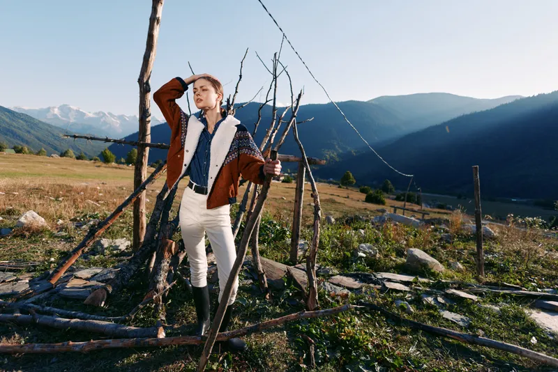 Woman in jacket and boots stands by a wooden fence in a countryside field with mountains in the background, posing for a fashion portrait amid open nature and sunlight.